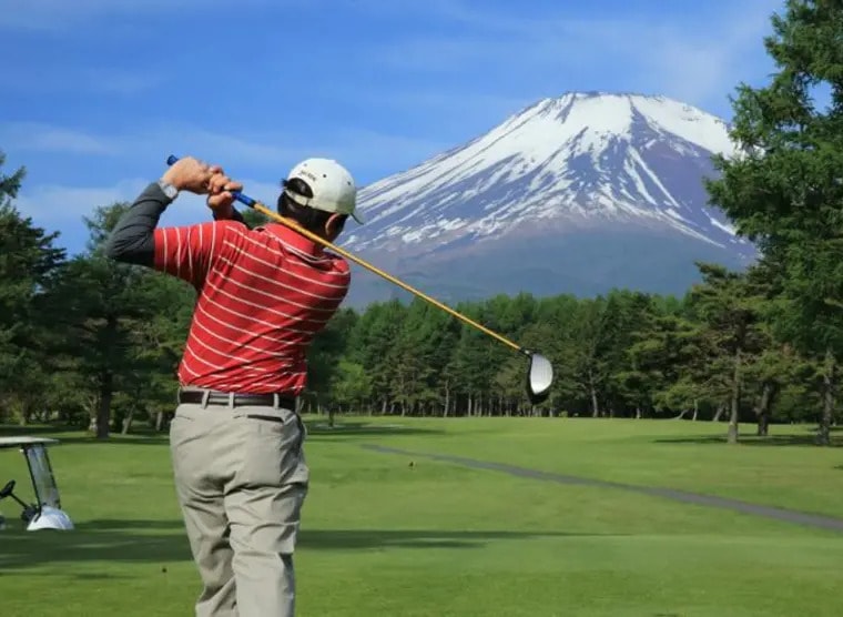 Golfer mid-swing on a Japanese course tee box with snow-capped Mt. Fuji rising above the tree line behind him