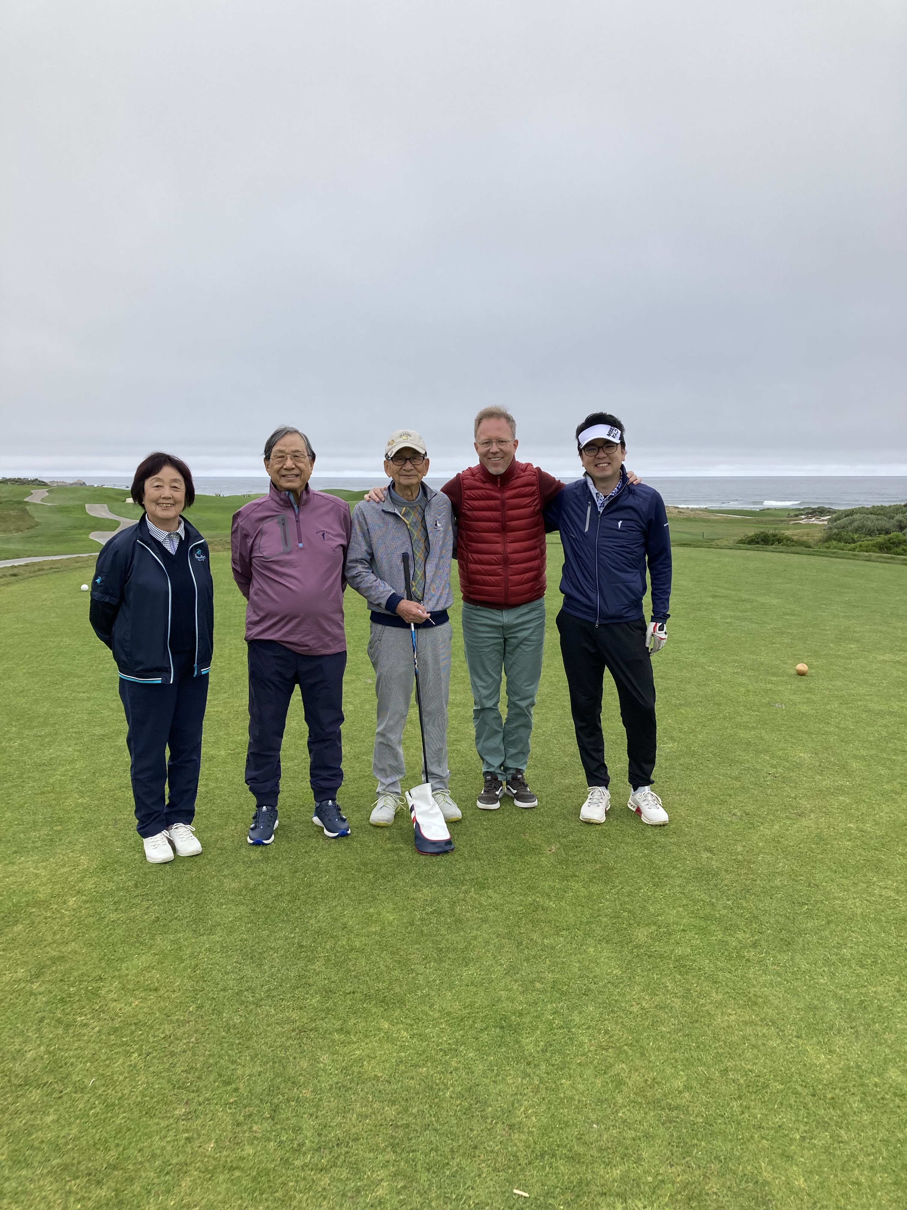 Five golfers posing on a coastal links course with a dramatic overcast sky and ocean views behind them
