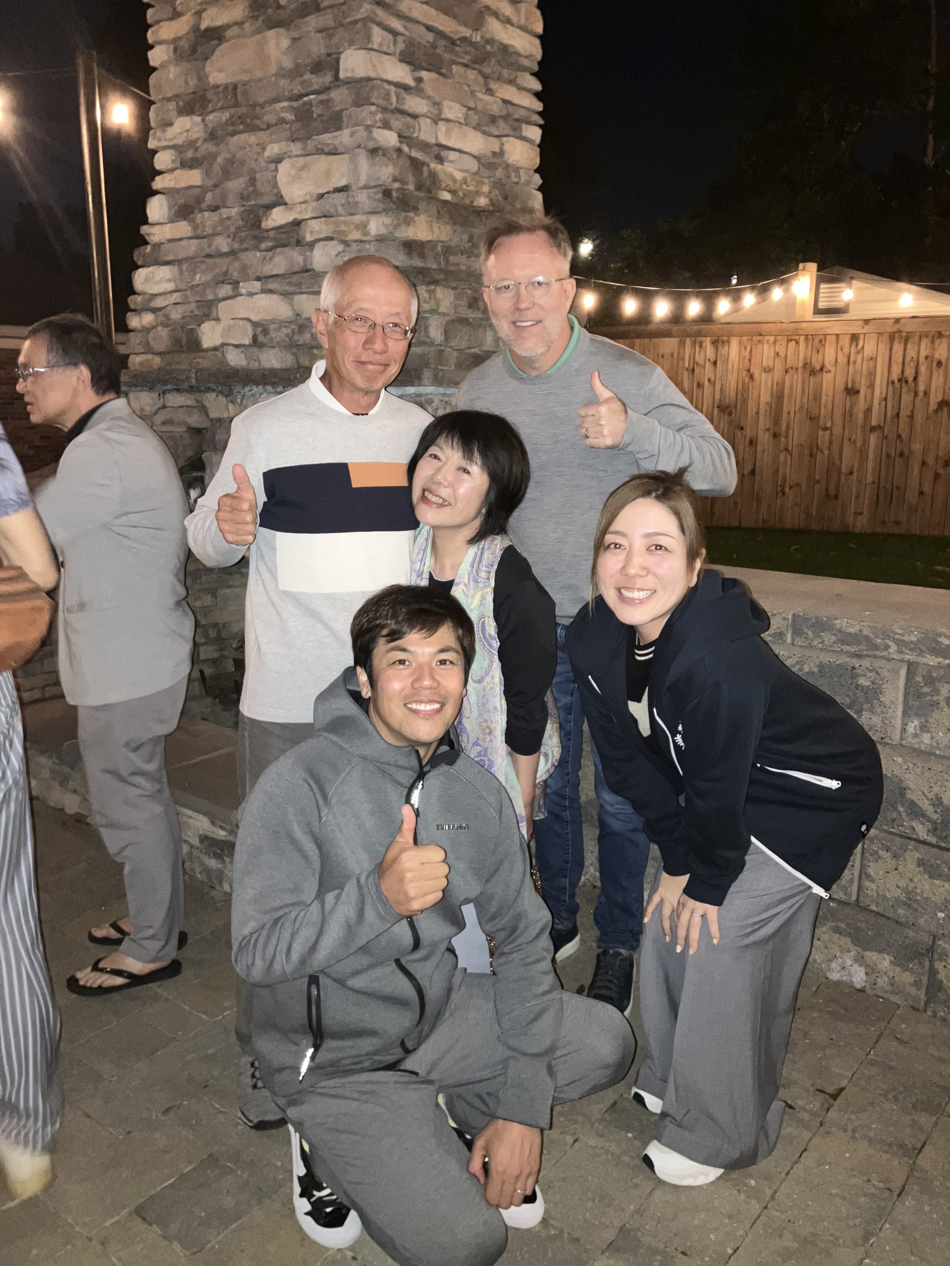 Mixed group of golfers and friends celebrating at an outdoor evening gathering with string lights and a stone fireplace