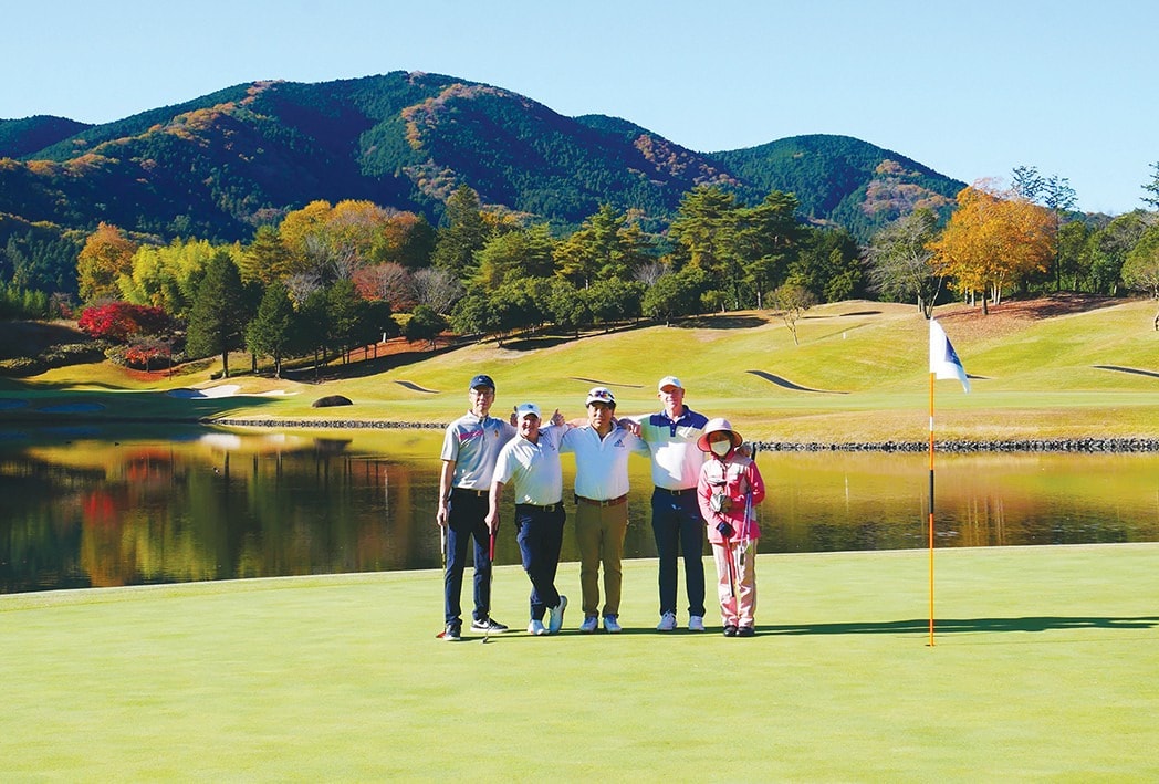 Fivesome posing on a Japanese golf course green in autumn, with a scenic lake and vibrant fall foliage in the background