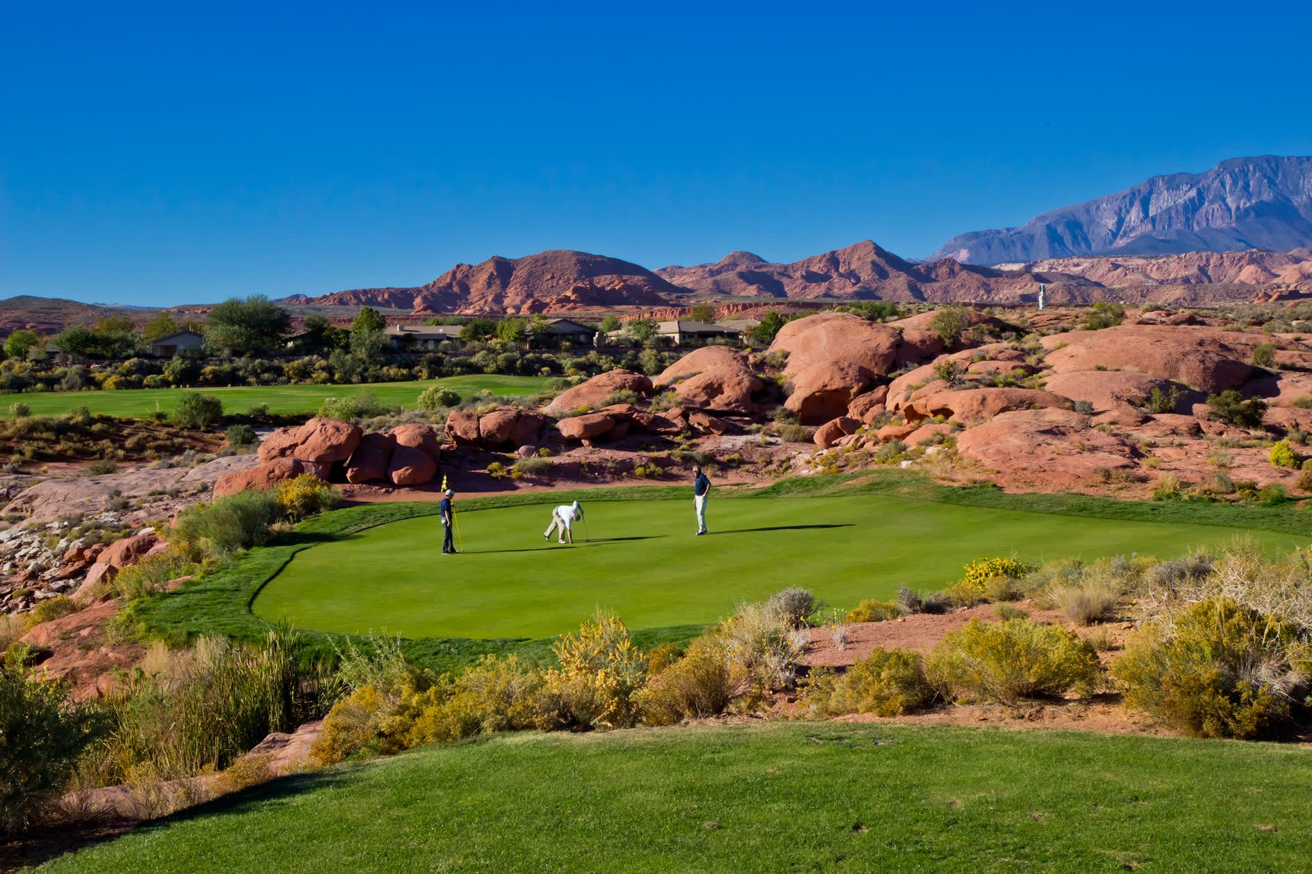 Three golfers on a putting green at a Utah desert course surrounded by dramatic red sandstone boulders and mountains
